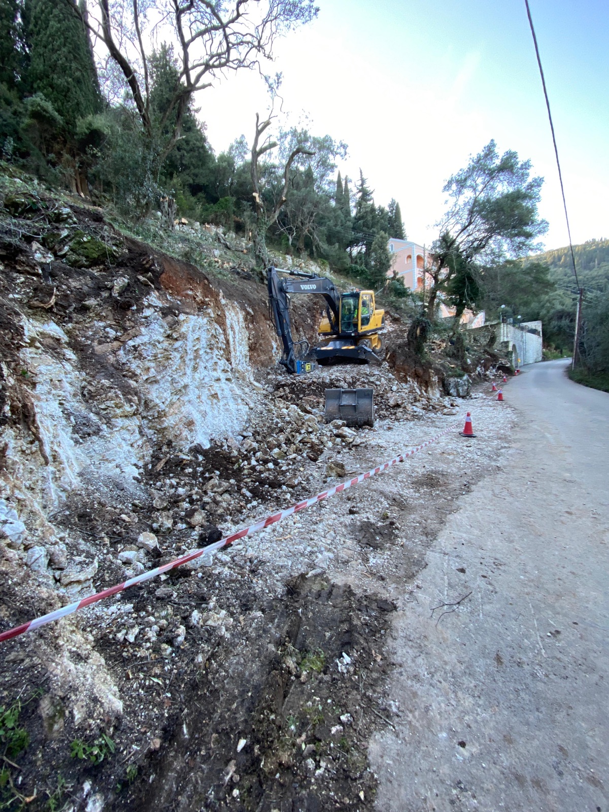 An excavator digging into the white rock face beside the narrow village road in fading dusk light.