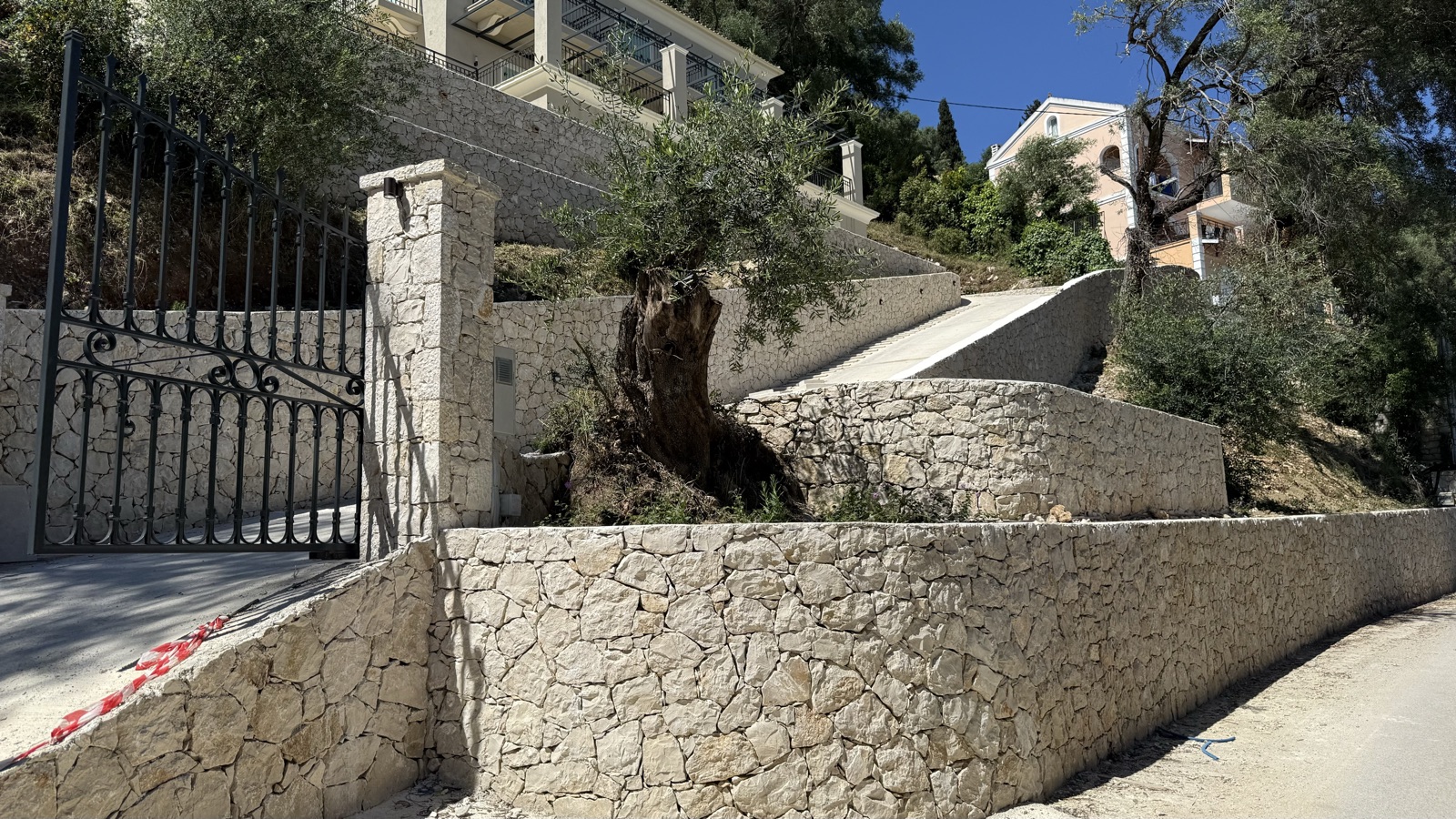 A black wrought-iron gate hung on a cut-stone pier, an ancient gnarled olive tree preserved within the curved limestone walls.