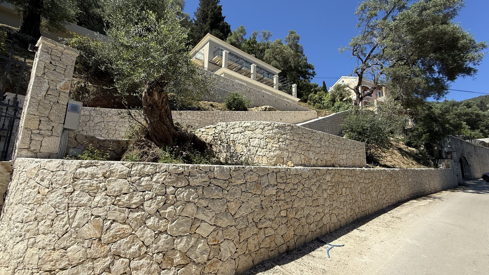 Tiered curved limestone retaining walls seen from the road, an olive tree on the upper terrace and the villa's glass balustrade at top.