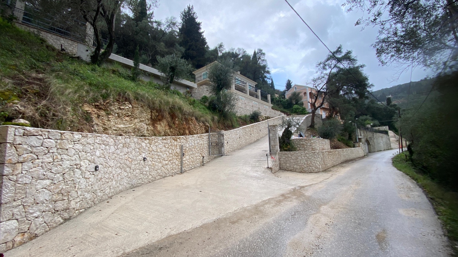 The completed limestone walls along the road on an overcast day, the open gate and driveway curving up to the villa.
