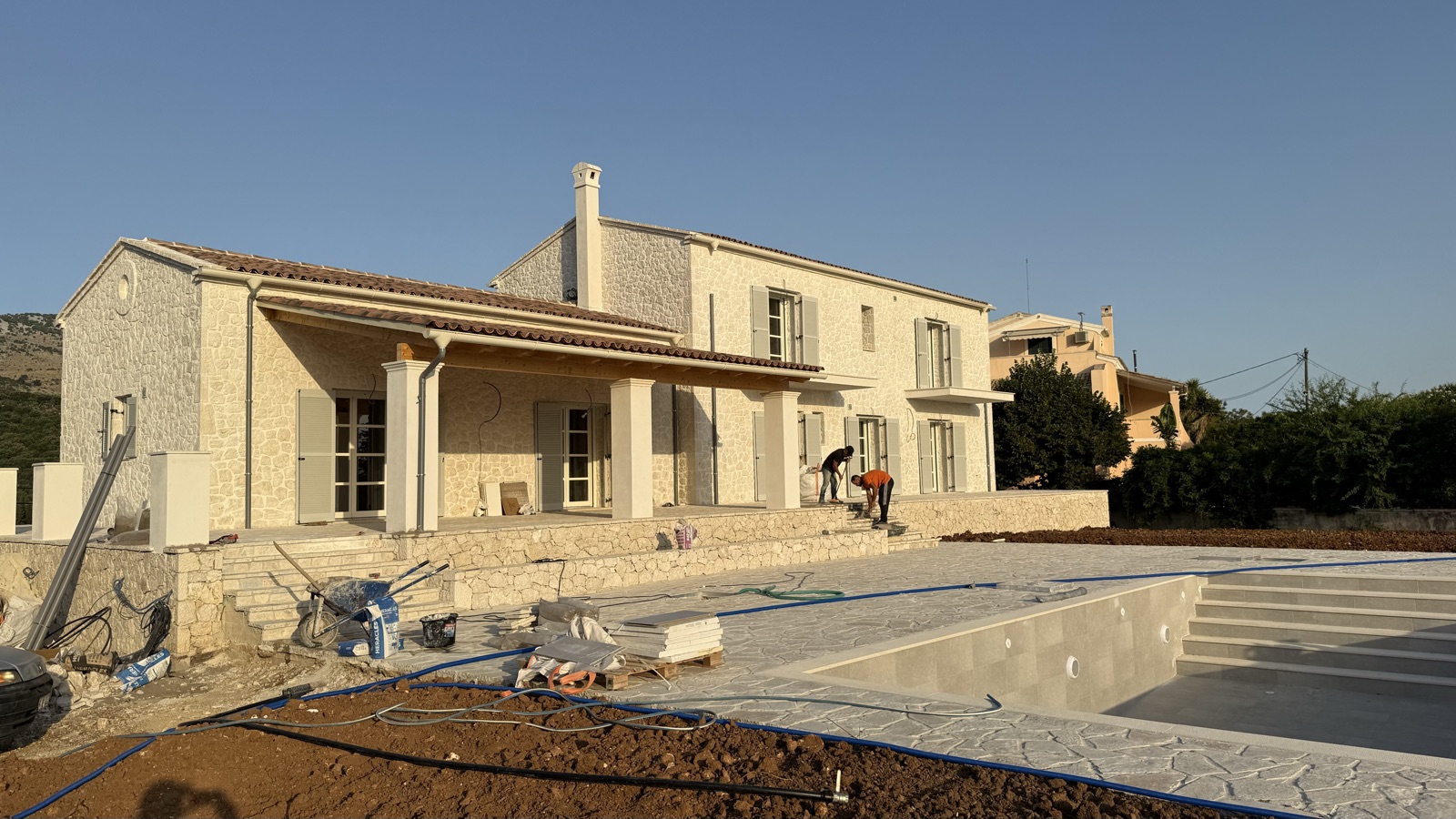 Cream stone-clad facades and a columned veranda at golden hour, the empty pool shell and stone paving in the foreground.