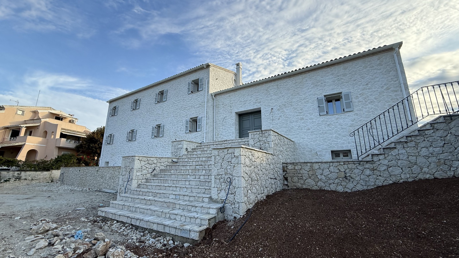 White-pointed stone facade with a grand exterior stone staircase and curved wrought-iron railing climbing to an upper terrace, stone retaining wall in the foreg