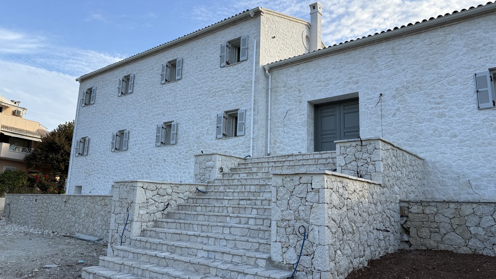Wide hand-laid stone steps between clad parapet walls leading to grey double doors, shuttered windows across the white-pointed limestone facade, even soft light