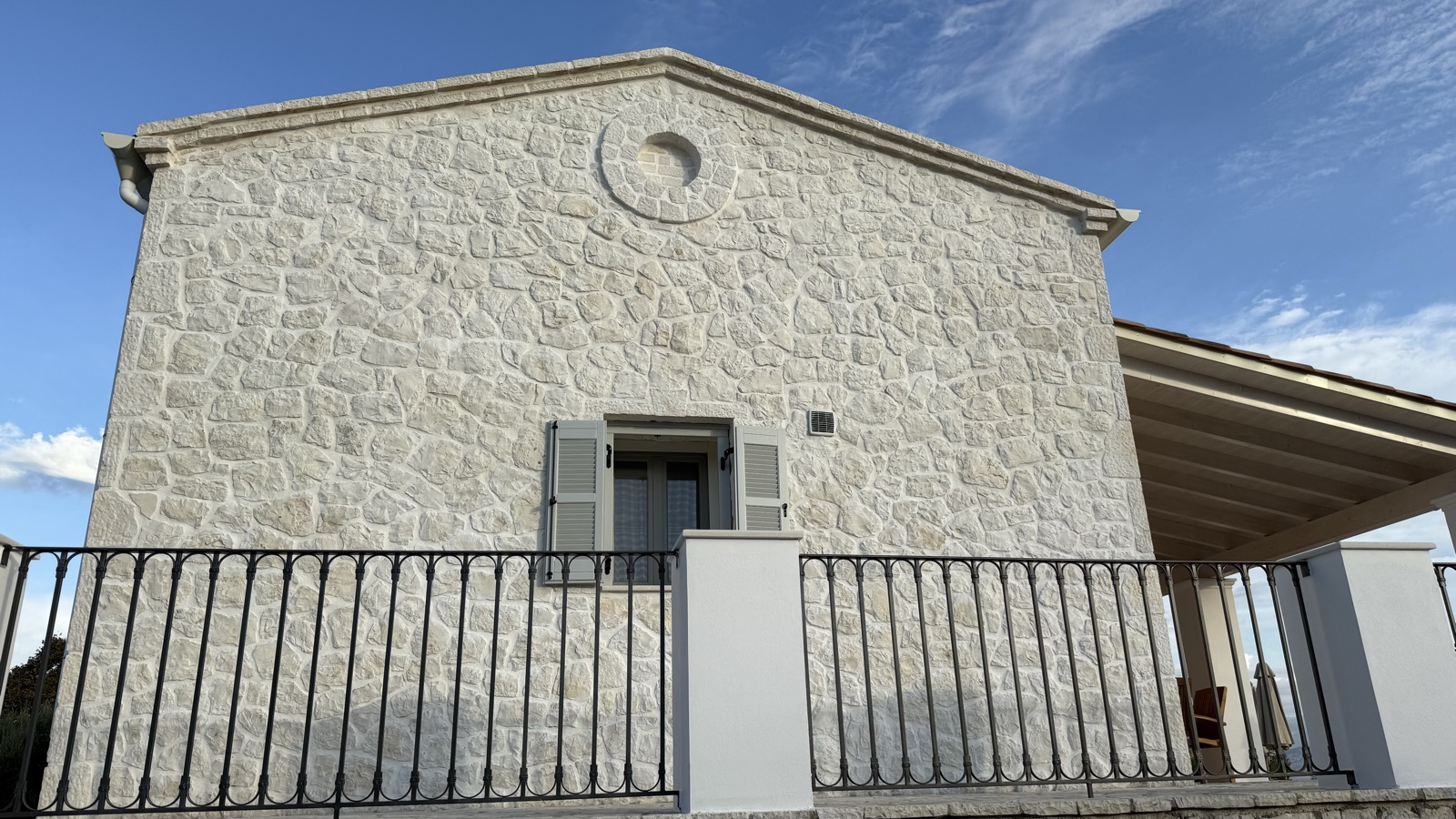 Rough cream limestone cladding with bright pointing, a circular stone oculus motif set into the peak, blue-grey shutters, and a black wrought-iron balcony rail 
