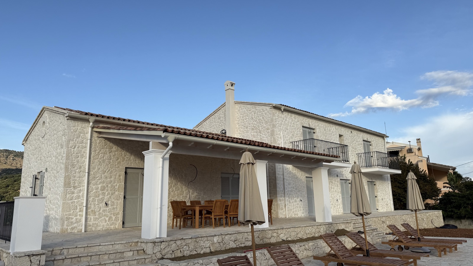 Stone columns and timber-look pergola roof with terracotta tiles, outdoor dining table, closed umbrellas, loungers on the lower terrace, wrought-iron balconies,